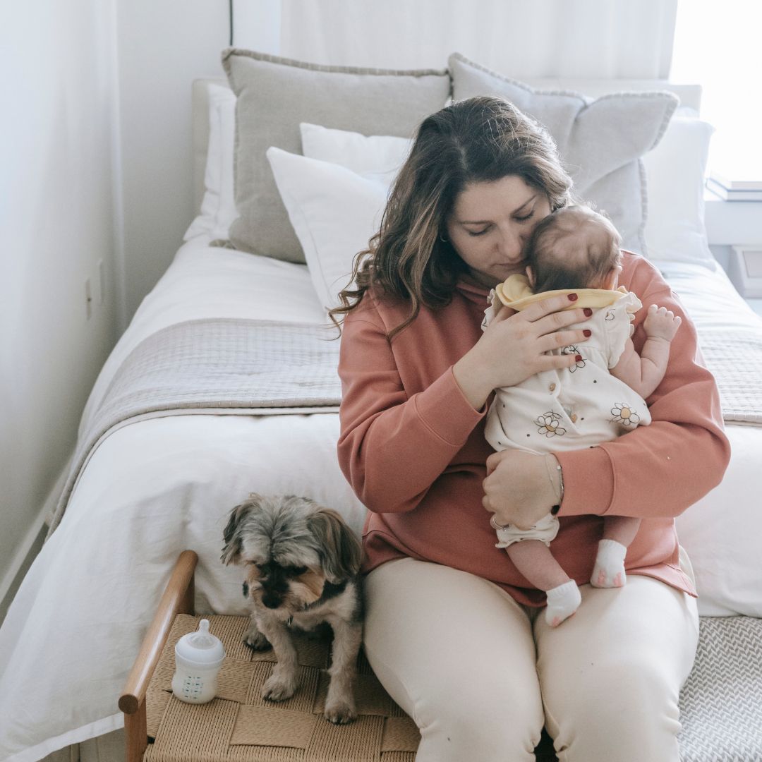A woman snuggles her little baby while the dog sits next to her.